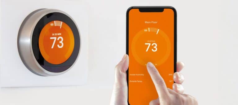 smart thermostat positioned on the wall with the phone that controls it being held in front of it being adjusted by the hands that are holding it