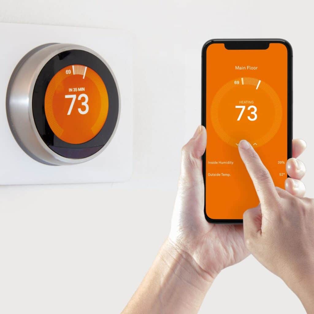 smart thermostat positioned on the wall with the phone that controls it being held in front of it being adjusted by the hands that are holding it
