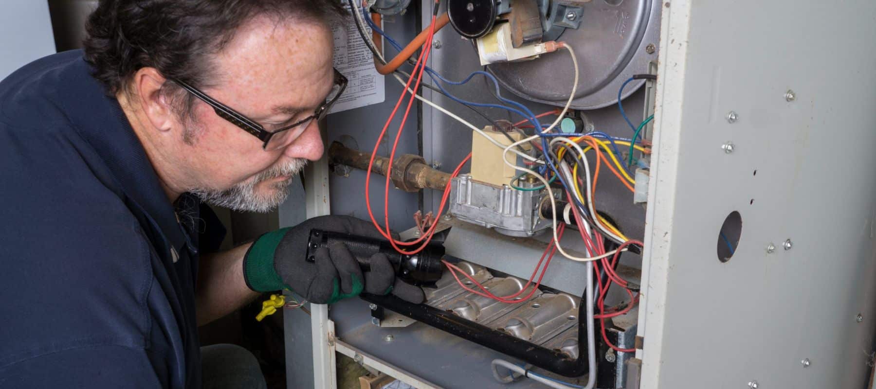 hvac technician using a flashlight to look inside a furnace to perform repairs