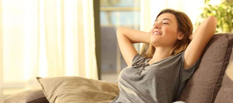 young woman sitting on a couch leaning back with her arms behind her head showing she's relaxed thanks to her cooled home