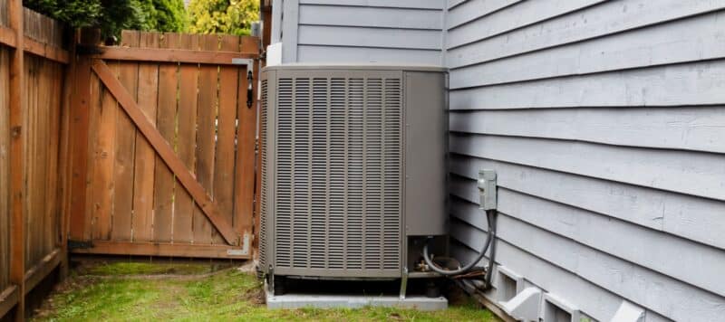 A heat pump unit installed beside the exterior wall of a house near a wooden fence