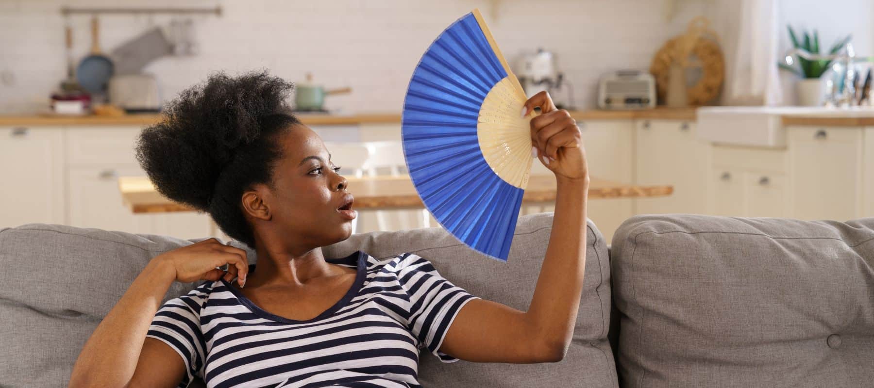 A woman sitting on a couch in a warm home fanning herself with a blue hand fan, looking uncomfortable due to the heat