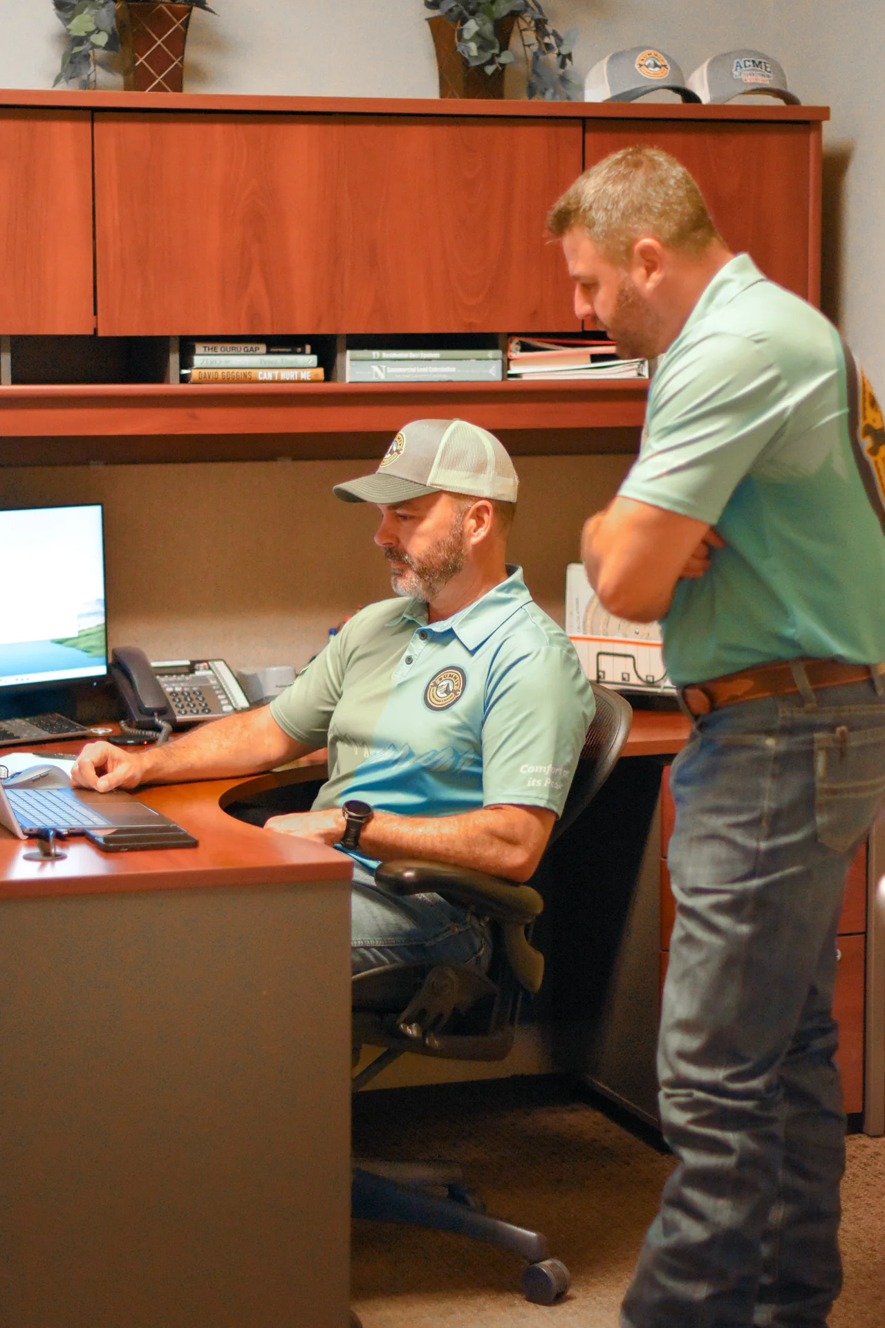 Two men in matching Summit uniforms discuss work at a desk with a computer in an office setting.