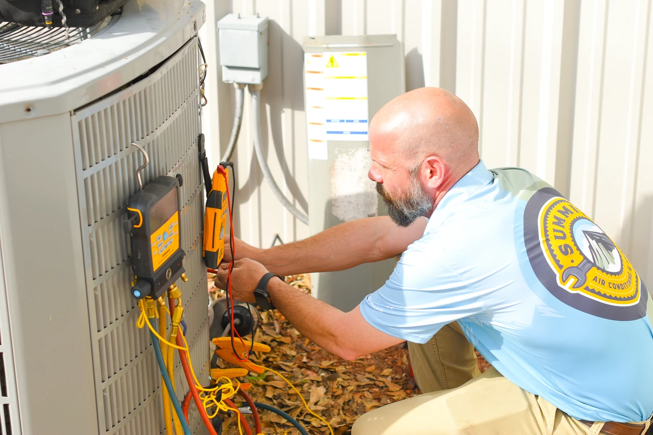 Summit technician in a blue shirt servicing an outdoor air conditioning unit with tools and gauges attached.