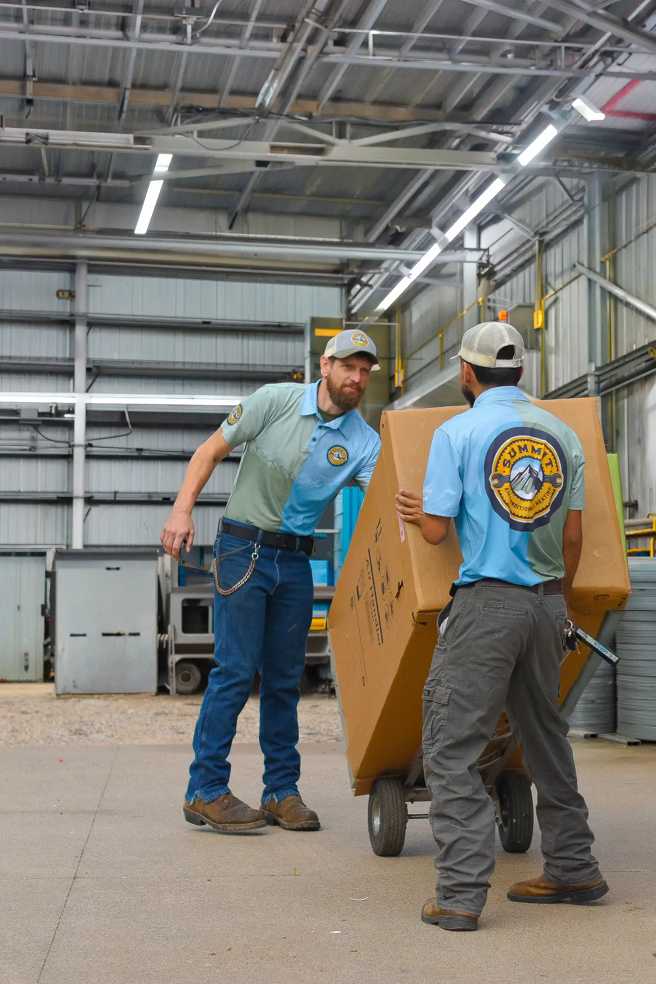 Two workers in Summit uniforms move a large box on a dolly inside an industrial warehouse.